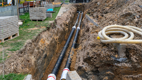 Open construction trench with black utility pipes, coiled protective conduit, paving blocks and excavated soil, showing underground infrastructure installation and urban road repair worksite.