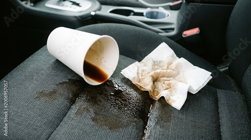 spilled coffee on car seat mess problem, Paper cup with spilled coffee lies on car seat with visible liquid stain on fabric. Used napkins nearby. Vehicle interior details in background. Mess, accident