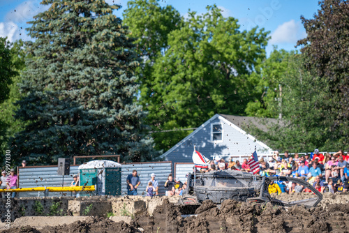 Crowd watches dirt bike event with mud flying in the air on a sunny day in a park setting