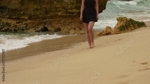 Woman walk along sandy shoreline with waves gently wash ashore against rugged rocks. Marinha Beach, Algarve, Portugal