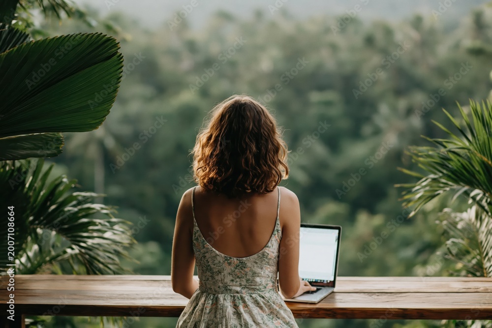 Fototapeta premium Woman in dress works on her laptop on wooden balcony surrounded by lush green plants with misty jungle landscape in background. High quality photo
