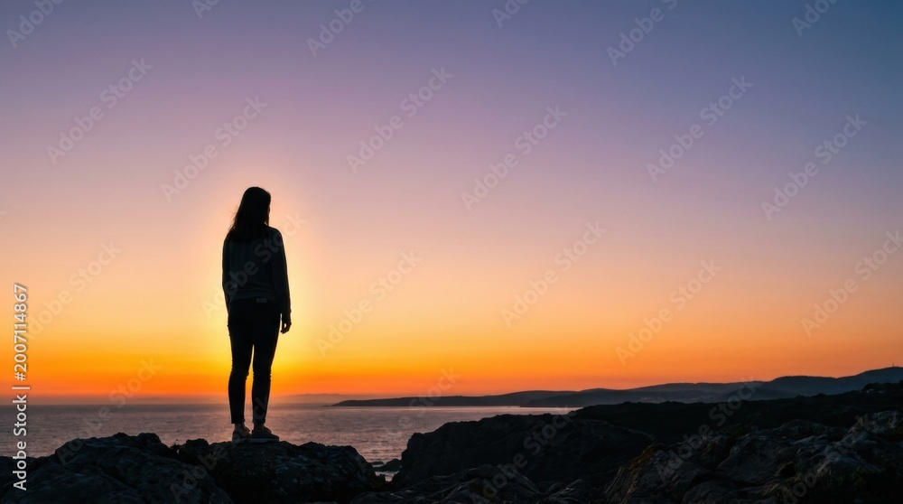 Obraz premium A person is standing on rocks by the sea during sunset. The sky shows bright colors of orange and blue. The scene captures the beauty of nature.