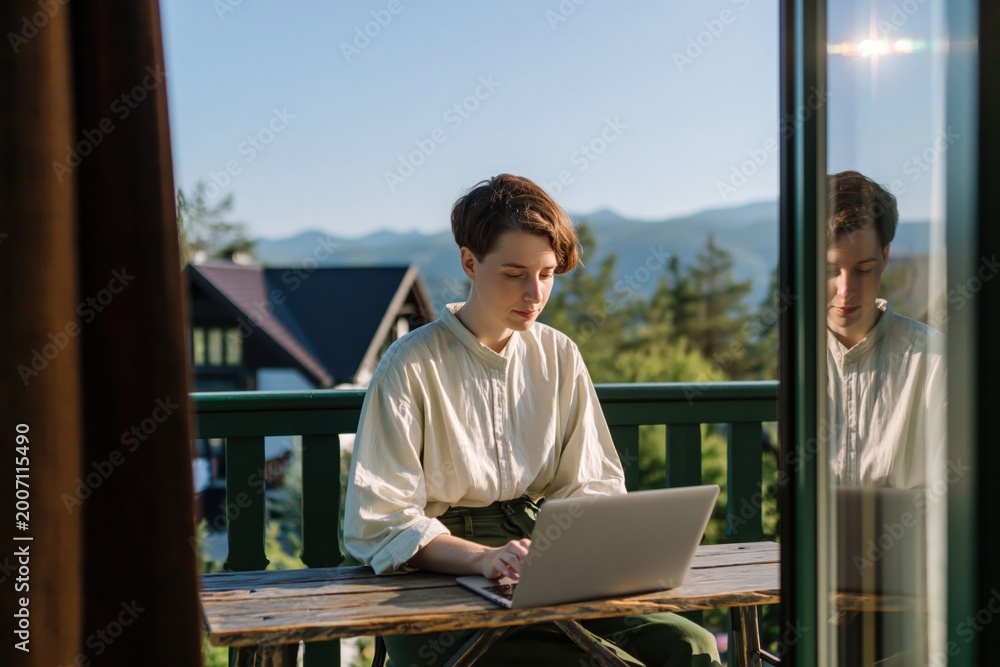 Obraz premium Woman is focused on her laptop while sitting on wooden balcony, surrounded by beautiful mountain landscape and clear blue sky. High quality photo
