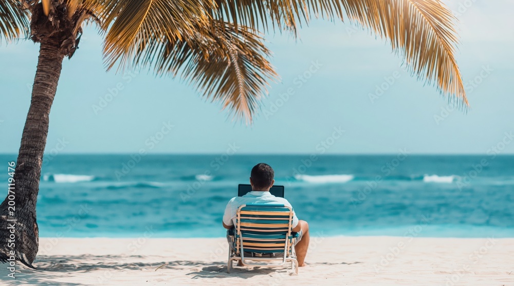 Fototapeta premium Man sits in striped beach chair, looking out at ocean from under shade of palm tree. High quality photo