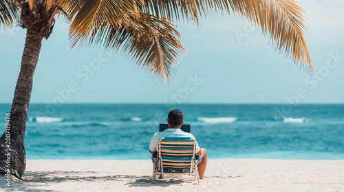 Man sits in striped beach chair, looking out at ocean from under shade of palm tree. High quality photo