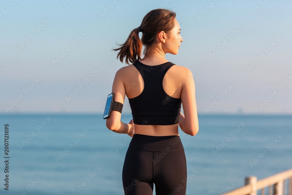 Naklejka premium Woman running by the ocean, focused on fitness and health, wearing sportswear and a smartphone armband at sunset