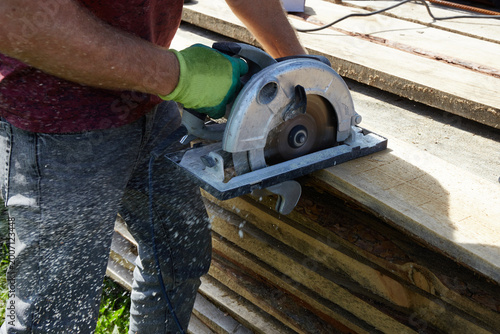 A worker saws wooden planks with a circular saw in a green, sunny place.