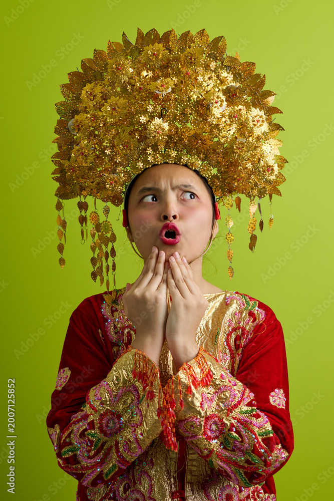 Fototapeta premium Surprised Young Indonesian Asian Woman Wearing Ornate Golden Headdress and Traditional Embroidered Costume Portrait