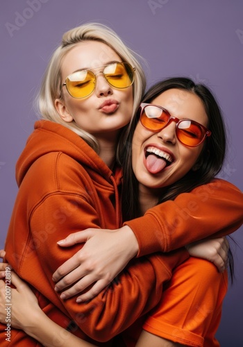 Two stylish young women friends wearing vibrant orange hoodies and colorful sunglasses posing for a fun studio portrait against a purple background