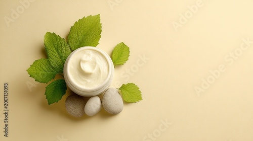 White cosmetic cream in a jar surrounded by fresh green mint leaves and smooth grey river stones on a beige background