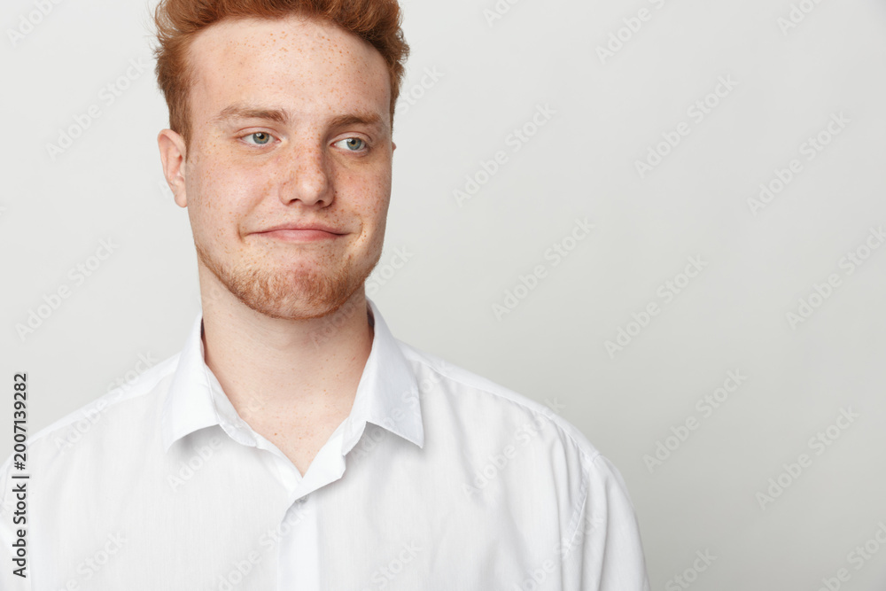 Obraz premium Young redhead man in white shirt looks sideways with skeptical expression on light background. Concept of doubt, uncertainty, and casual portrait of male model with freckles.