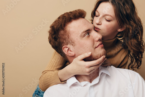 Young couple in casual clothes embraces tenderly on beige background. Woman holds man’s face gently, expressing love and intimacy. Concept of romance, affection, and close relationship.
