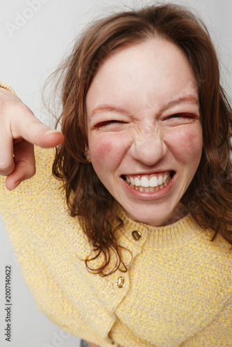 Happy young woman with curly hair wearing yellow sweater smiles widely and points finger at camera. Close-up portrait on light background. Concept of joy, fun, and positive emotions.