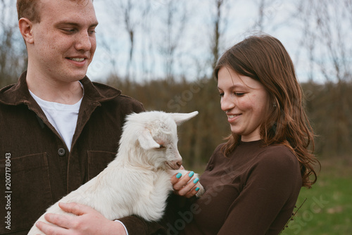 A young couple interacts with a small white baby goat outdoors during a casual lifestyle moment in early spring, smiling and enjoying the gentle animal together.