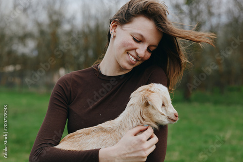 A woman warmly embraces a small goat kid in a green field, smiling with affection, enjoying a simple outdoor lifestyle, gentle moment between human and animal, peaceful rural scene.