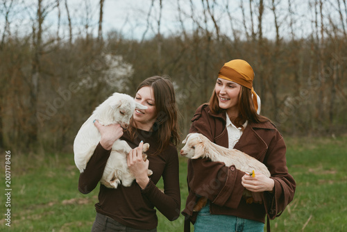 Goat goats kid animal animals lifestyle outdoor countryside women sharing a lighthearted moment with two kids held by friends in an open field, soft daylight, casual outfits, warm smiles,