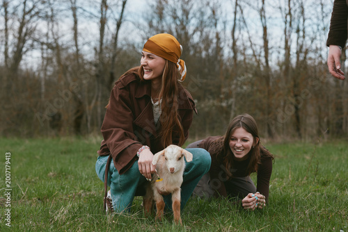 Two cheerful girls pet a curious goat kid in a grassy field, enjoying a carefree outdoor moment, natural smiles, warm clothing, gentle breeze, countryside vibes, and a playful, friendly mood.