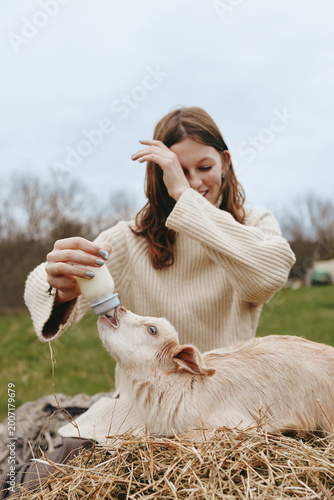 Young woman with long brown hair feeds a baby goat with a bottle while sitting outdoors on hay in a casual sweater, enjoying a peaceful lifestyle moment on a cloudy day.