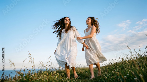 Two happy young women in white dresses walking barefoot in a grassy field under a blue sky, enjoying nature and freedom.