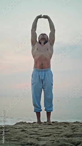 Man Stretches Arms Upward On Sandy Beach. Calm Beach Scene With Man Practicing Mindful Stretching Exercises. Full Body Stretch By Asian Man During Peaceful Morning On Tranquil Seaside