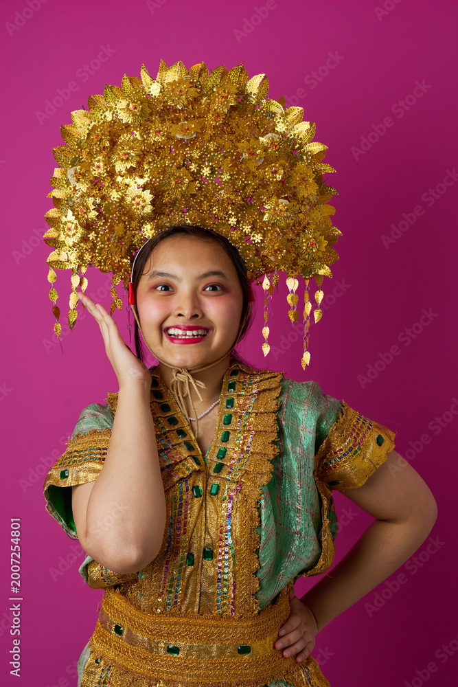 Fototapeta premium Smiling Indonesian Asian Girl Wearing Ornate Gold Headdress and Traditional Costume Against Pink Background