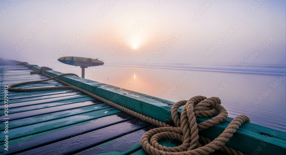 Naklejka premium Nautical ropes resting on a wooden pier overlooking a misty beach at sunrise. Soft morning light illuminates the fog over calm ocean waves. Coastal serenity concept, ideal for travel marketing.