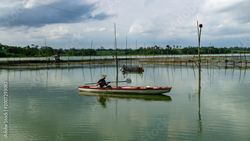 Tableau sur toile A lone fisherman fishing from a small wooden boat on a calm lake, East Java