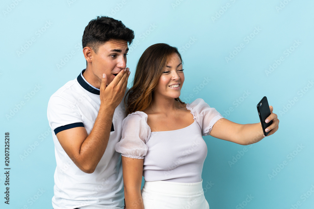 Naklejka premium Young caucasian couple isolated on blue background making a selfie with the mobile
