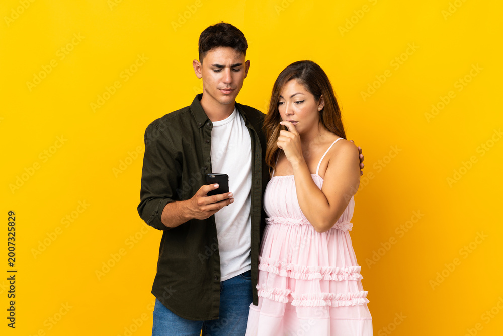 Naklejka premium Young caucasian couple isolated on yellow background reading a message with the mobile
