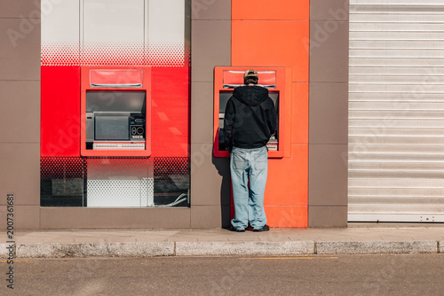 Billede på lærred Young man in black hoodie and baggy jeans withdrawing cash at a red ATM on a sun