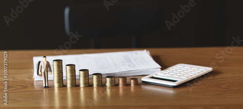 Stacked coins with a miniature businessman and calculator on a wooden desk, symbolizing financial growth, investment planning, and business success