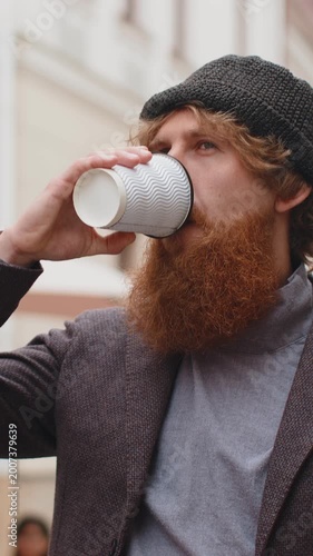 Joyful young man enjoying morning coffee or tea hot drink outdoors. Relaxing, taking a break. Redhead bearded boy walking in urban city center street, drinking coffee to go. Town lifestyles outside