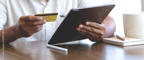 Person holding credit card and using tablet for online shopping at home, with coffee and notebook on wooden desk, ideal for e-commerce, digital payment, fintech, and modern lifestyle concepts