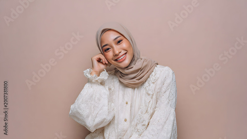Beautiful young Asian Muslim woman in hijab smiling and leaning on hand against beige background