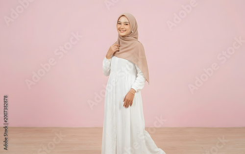 Beautiful young Asian Muslim woman in hijab smiling and leaning on hand against beige background