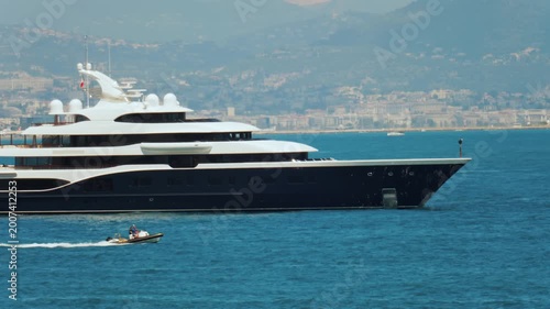 Large luxurious superyacht anchored in blue waters, small boat speeding by, picturesque coastal town.