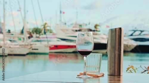 Red wine glass sitting on table by harbor with luxurious yachts docked in background during sunny day.
