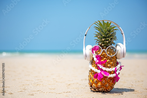 Summer in the party.  Hipster Pineapple Fashion in sunglass and listen music with sunblock and sandal on the sand beach beautiful blue sky background.  Fashion Summer Vacation Concept, copy space