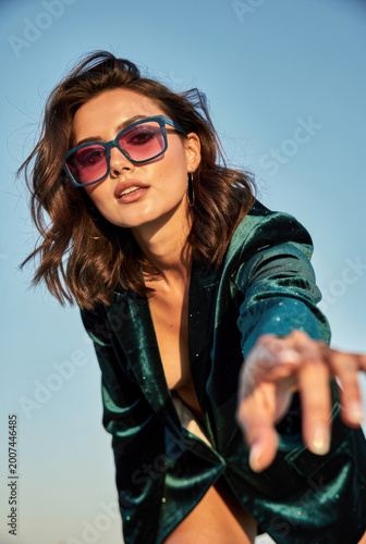 Confident Fashion Portrait of Woman in emerald velvet Outfit with Red Sunglasses gesturing towards Camera