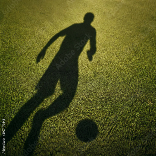 dynamic soccer player shadow dribbling on green grass pitch long late afternoon sunlight ball shadow separate abstract athlete motion no visible player aerial top down view striking contrast sports co