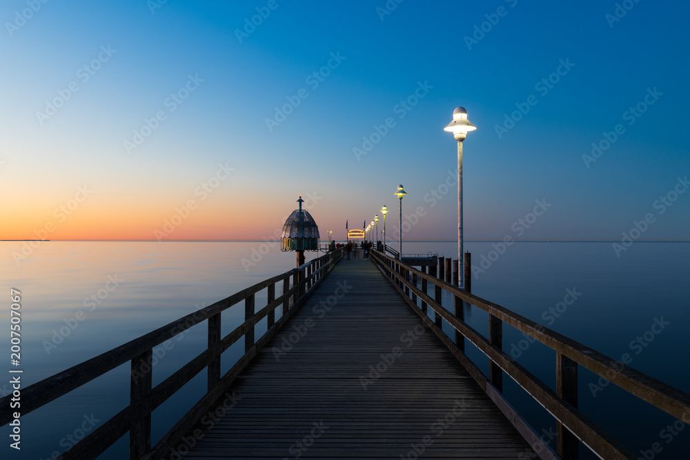 Fototapeta premium Seebrücke Zinnowitz bei ruhiger See zur Dämmerung mit Holzsteg und Lampen über der Ostsee unter klarem Himmel