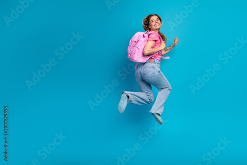 Fotografie Young woman with pink backpack jumps in the air against a blue background wearin