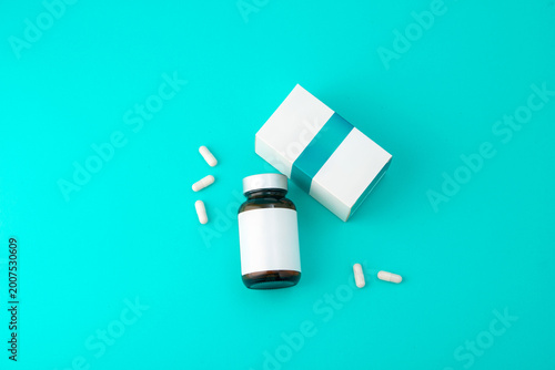 An amber glass medicine bottle with a blank white label is placed on a turquoise background next to its packaging box and several white capsules.