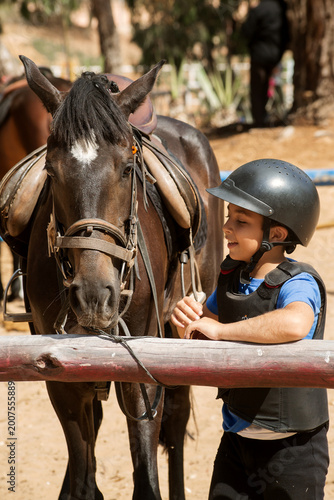 A young boy wearing a protective helmet and riding vest stands beside a saddled horse at a riding stable. 