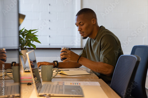 African American adult man sitting at meeting table holding mug, working on laptops and notebook