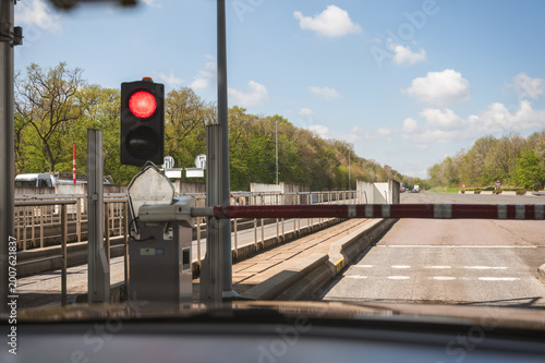 Red traffic signal and closed barrier at toll booth entrance of paid highway, green trees and blue sky in background, road safety checkpoint and European motorway infrastructure concept