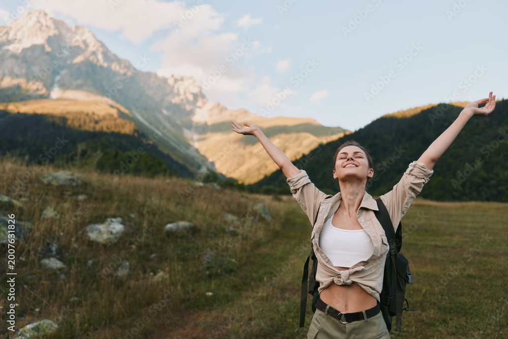 Naklejka premium Woman traveler with backpack raises arms in triumph in a meadow beneath mountains, enjoying fresh air and sunlight, embracing nature and outdoor adventure in a carefree lifestyle