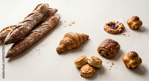 Freshly baked artisan bread including baguettes, croissant, danish pastry, and assorted rolls arranged on a light surface with crumbs scattered around