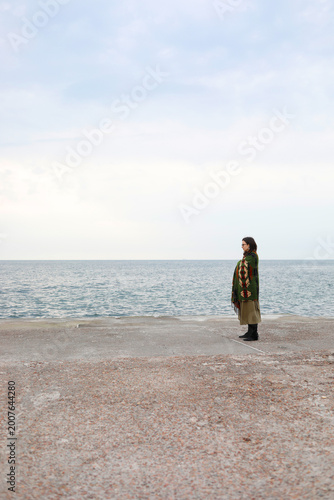 Man in green jacket stands alone on a wide pier facing the sea under cloudy sky, open space and muted tones creating a calm, minimal and contemplative coastal scene