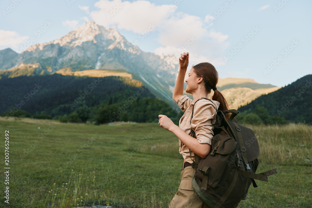 Naklejka premium Cheerful woman hiking outdoors, raising a fist in celebration, wearing a backpack, in a lush valley with distant mountains, bright sky and natural colors